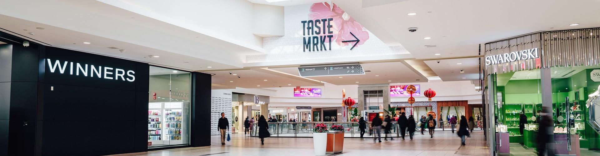 Shoppers walking through a shopping centre corridor past Winners and Swarovski stores with displays