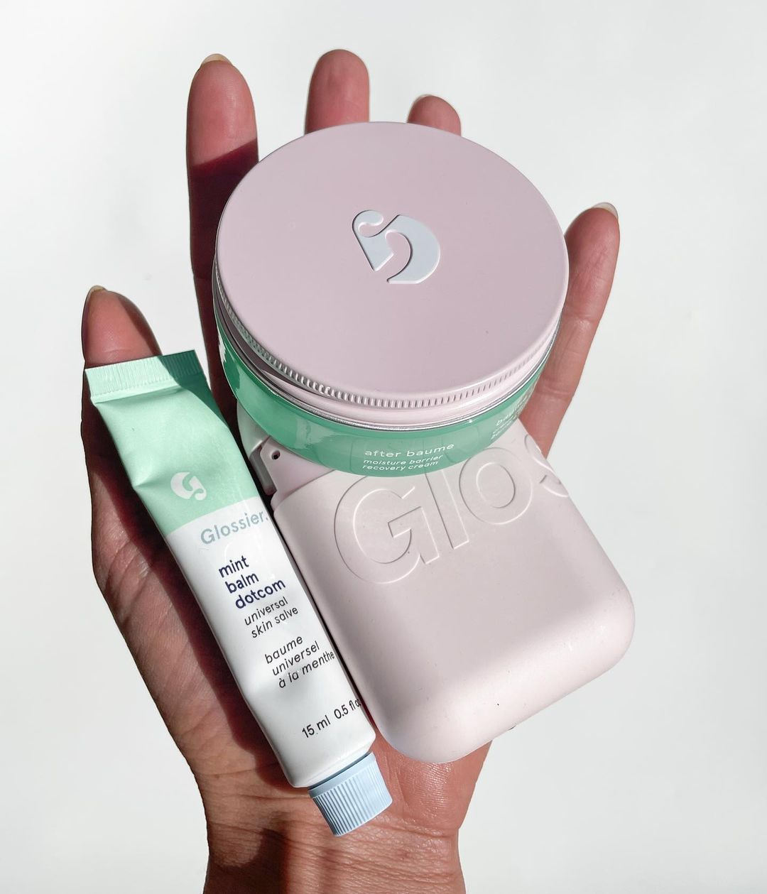 There is a hand holding up three products. The first, on the right, is a lip balm in a long squeeze tube container. The top product is in a circular container with a pink lid. And then bottom product is a rectangular pink container with a rounded edge.