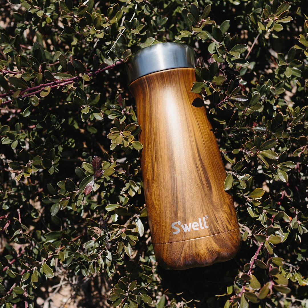 A brown textured water bottle with silver lid lying in greenery.
