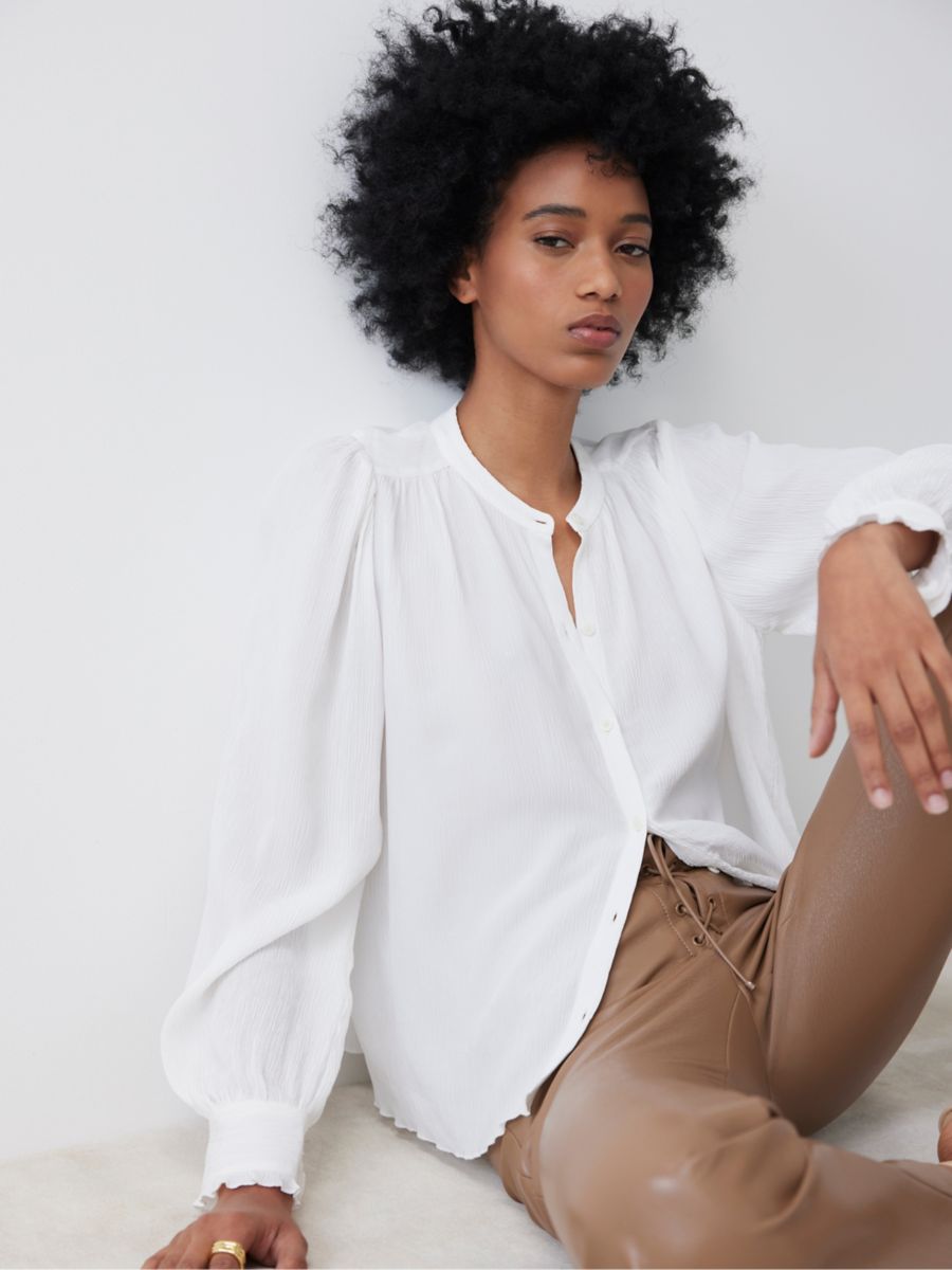 Model sitting against a white wall with her left leg bent up. She is wearing a white, loose blouse and light brown leather pants.