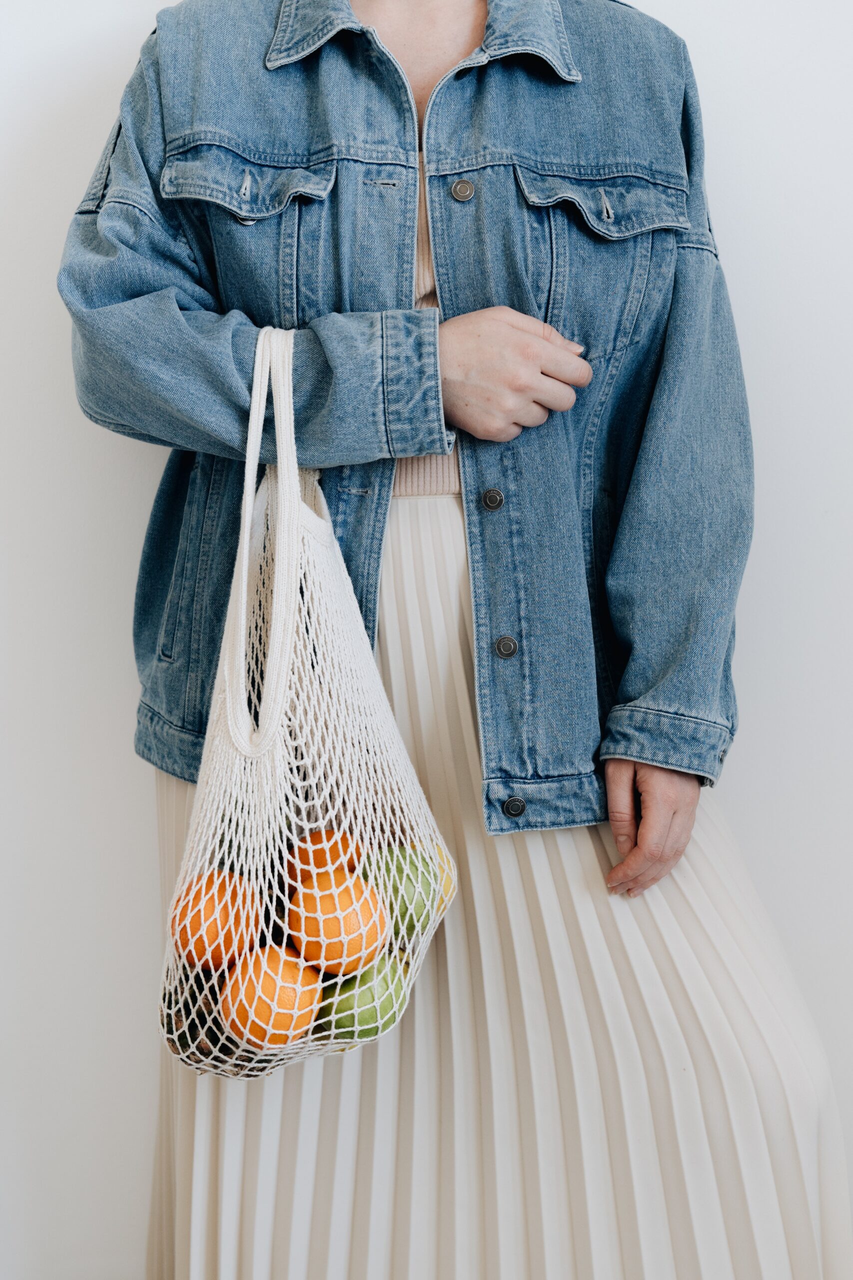 Person wearing a denim jacket holding a reusable bag with fruits