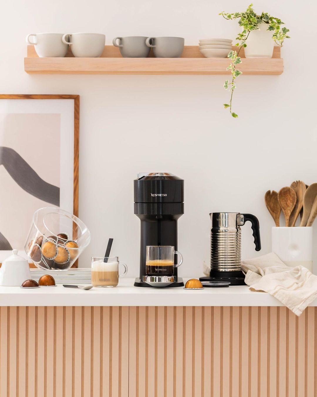 A ledge in the kitchen with a nespresso machine, frother, and container with wooden spoons.