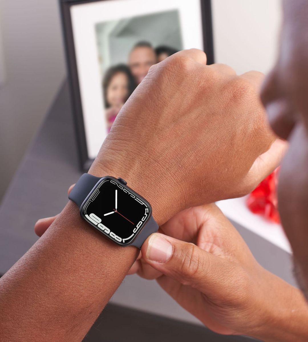 Close-up image of a person wearing a smart watch on his left wrist. There is a family photo in a frame in the background