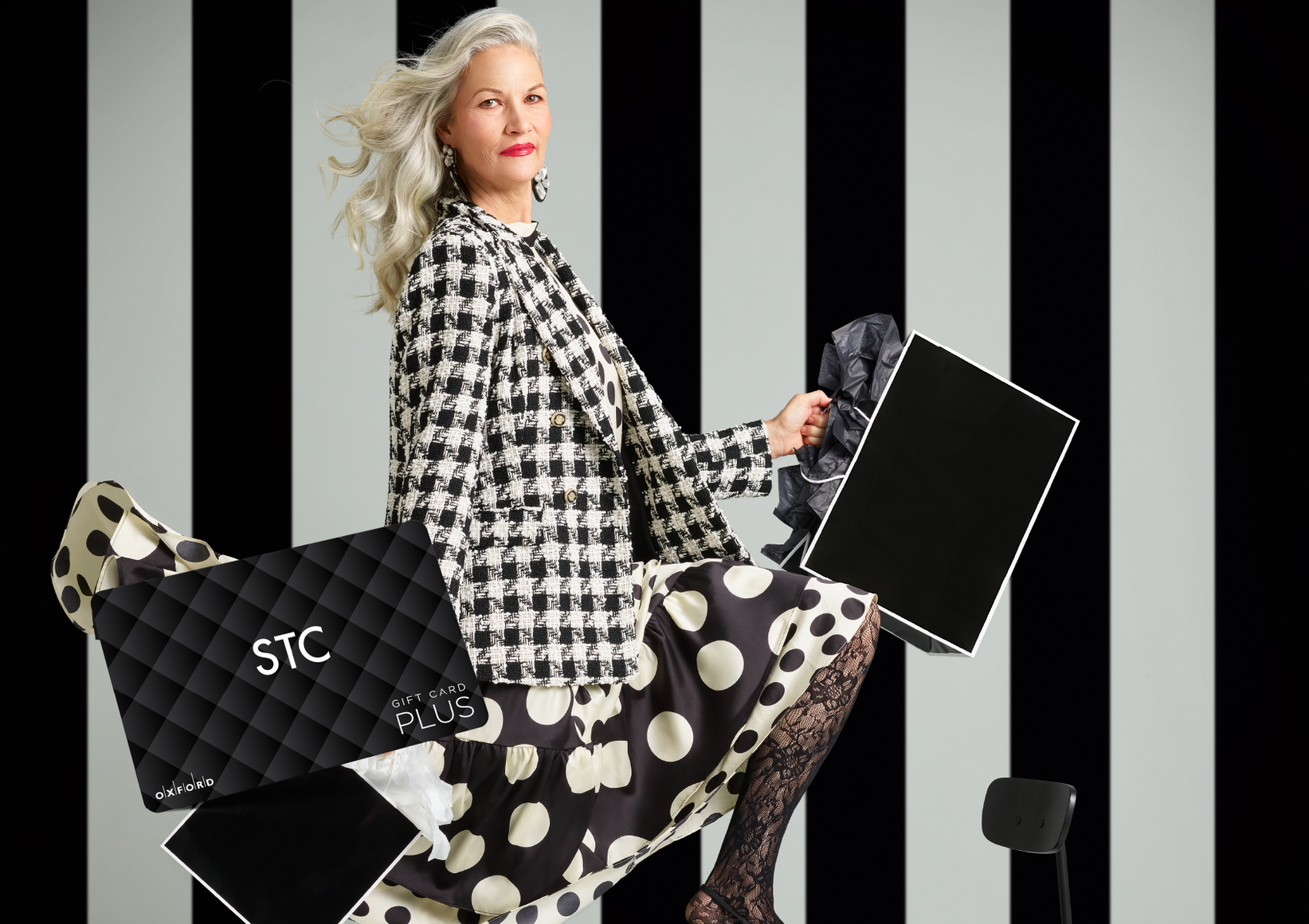 promotional image for a STC gift card. It shows a woman wearing a black and white houndstooth blazer and black and white polka dot skirt holding black and white shopping bags. A STC gift card is also in the image