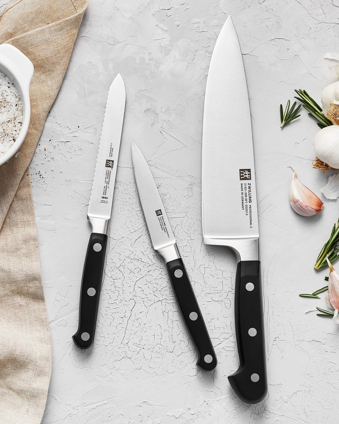 Three knives of different sizes sitting on a countertop with salt to the left and garlic gloves to the right.