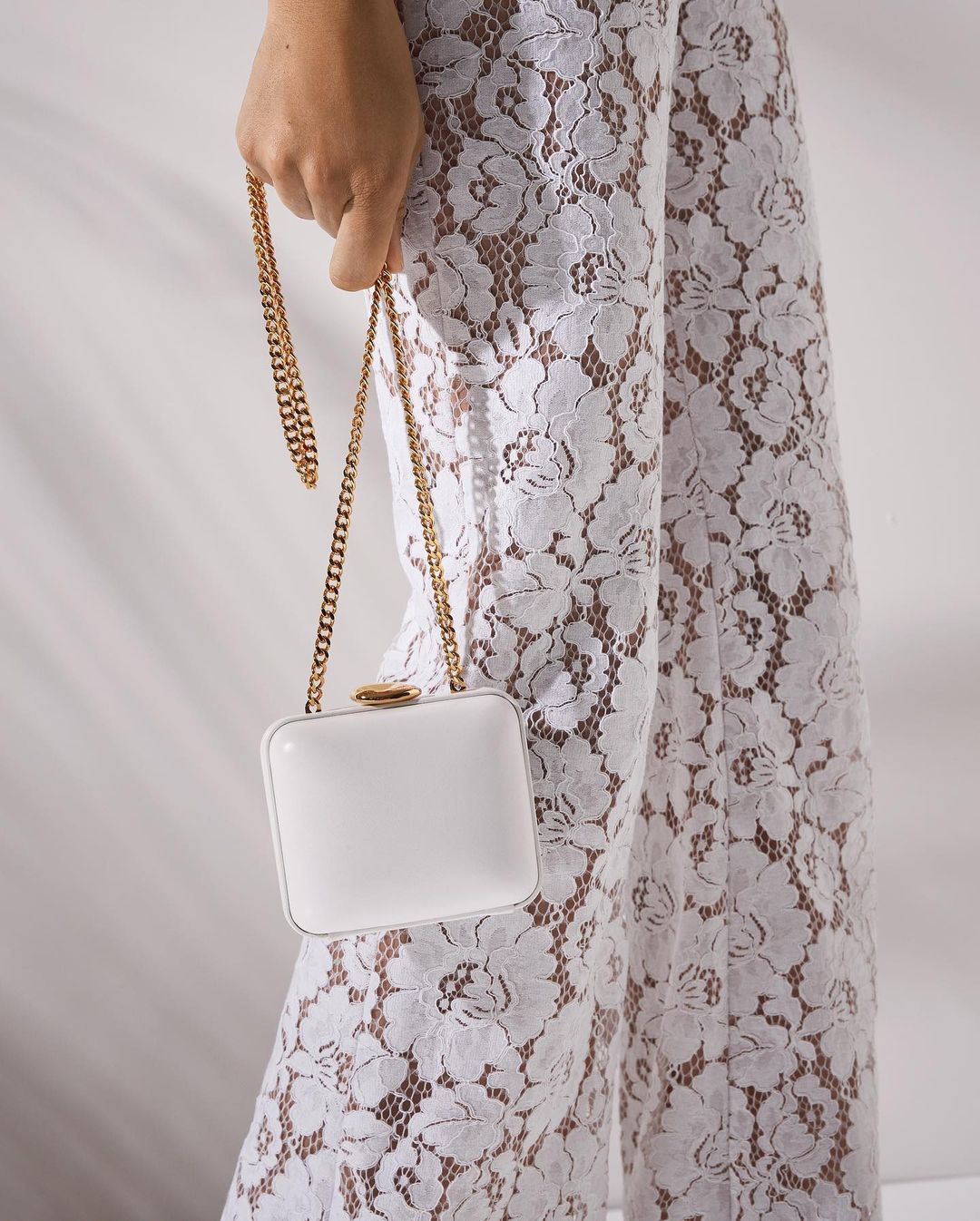 close-up image of a model wearing white lace pants and holding a white clutch with a gold chain