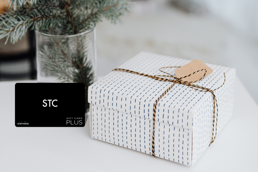 promotional image featuring a black STC gift card. beside the gift card is a present wrapped in white wrapping paper and behind the gift card is a vase with pine tree branches.