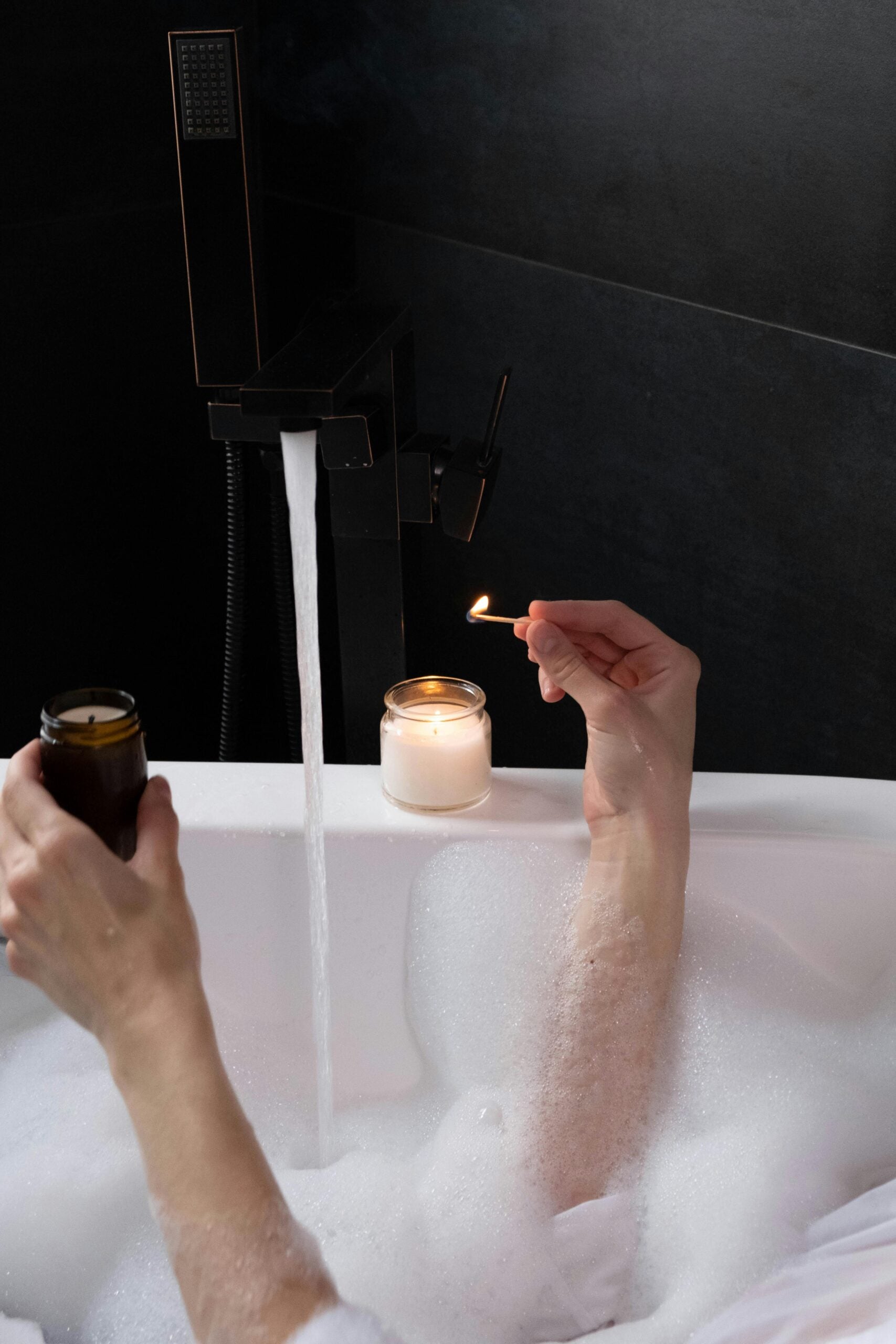 The arms of a person in a bubble bath, lighting a candle that sits on the edge