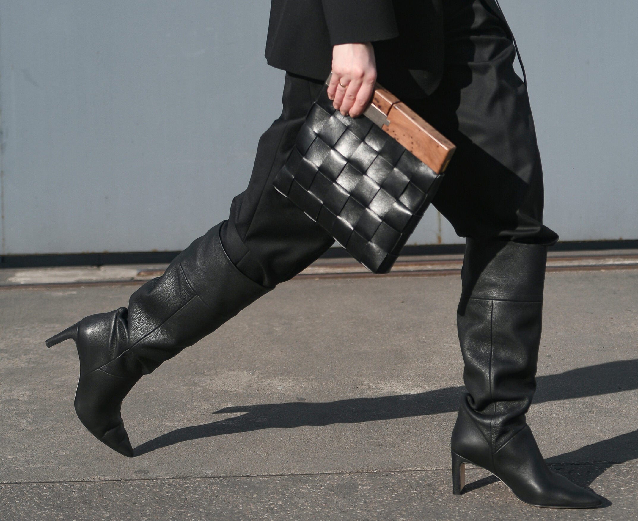 The legs of a person wearing tall black boots, carrying a black clutch.