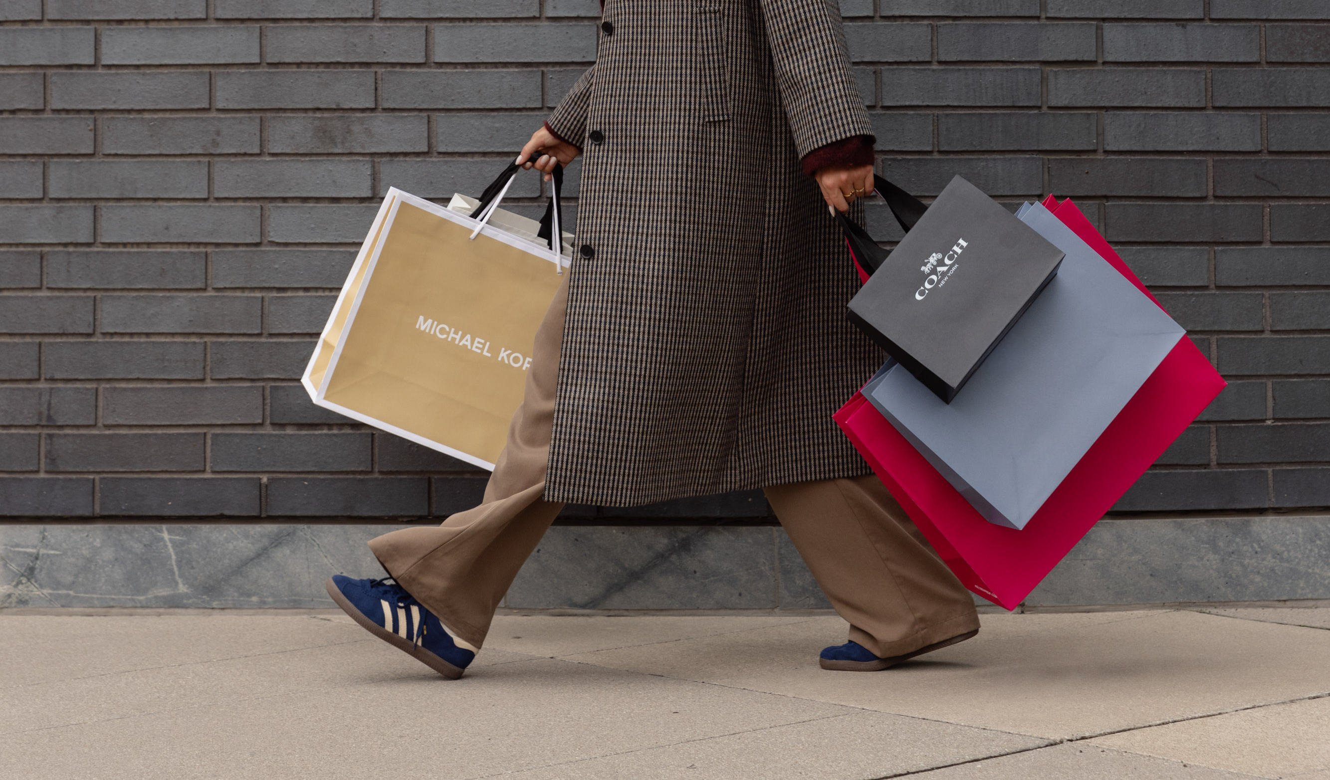 A woman in a trench coat walks with various shopping bags in both hands.